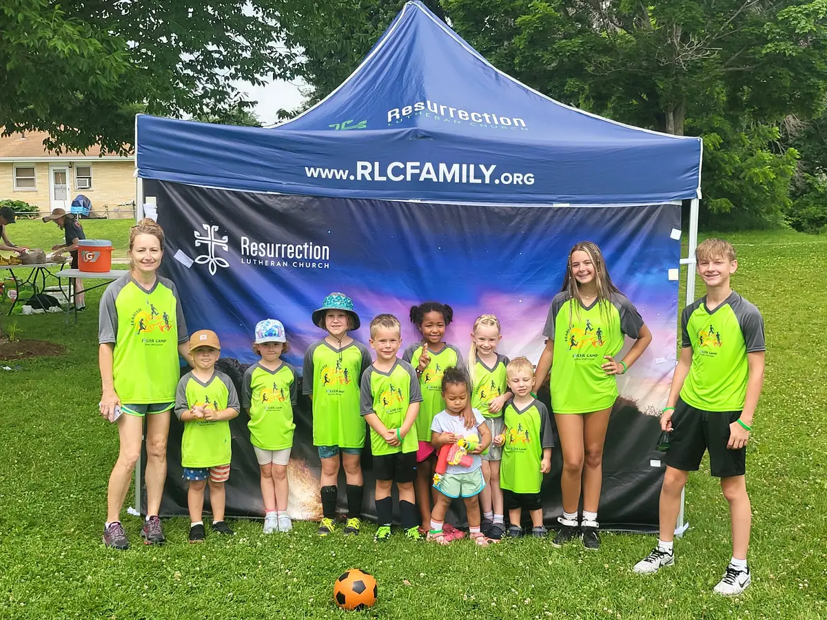 Children in soccer jerseys at a Resurrection Lutheran banner after soccer camp.